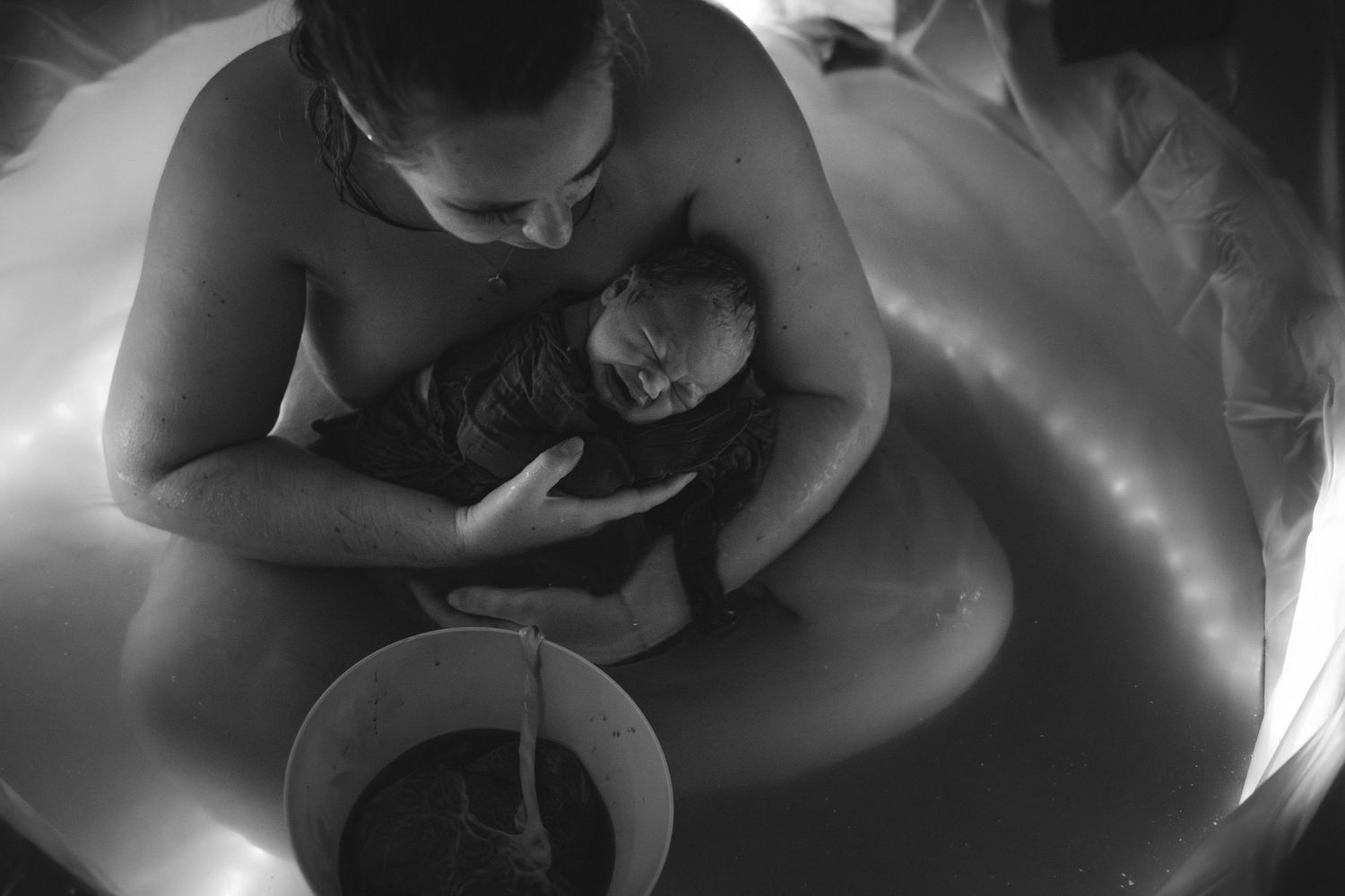vrouw houdt haar kindje in haar armen in het bevalbad waar zij net is bevallen. de placenta is nog verbonden aan de baby als een halve lotus