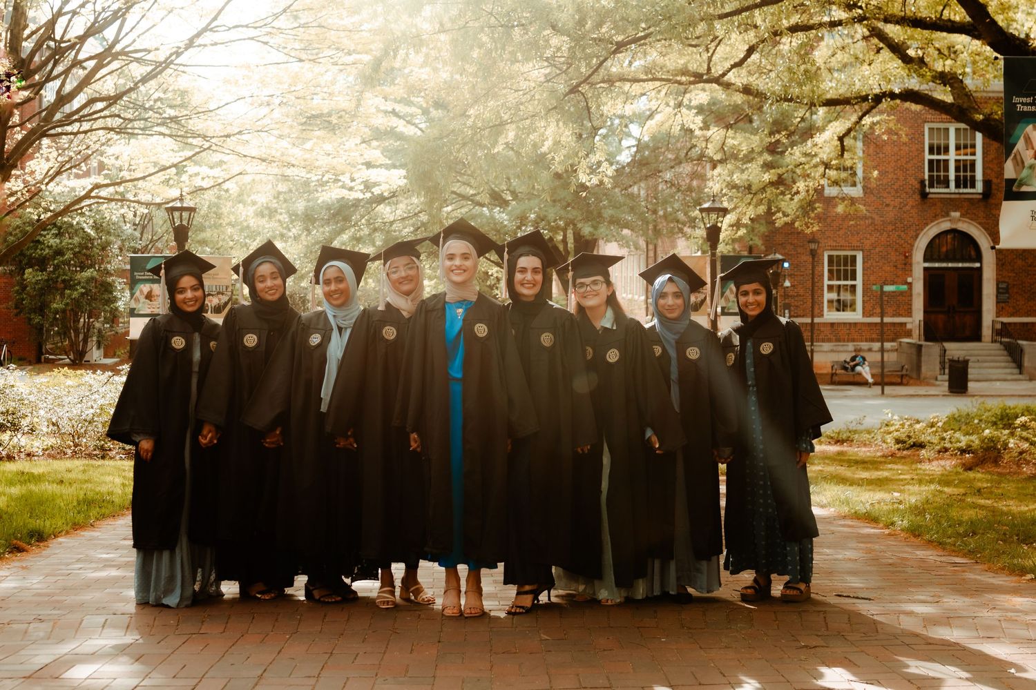 Group of graduates in black robes and caps standing together outside a brick building on graduation day.