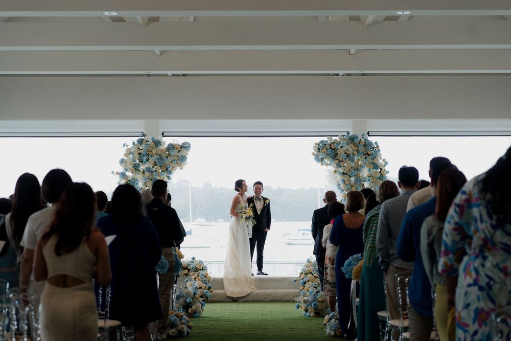 Elegant wedding ceremony with couple at floral arch altar, guests seated in aisle rows, overlooking scenic waterfront venue.