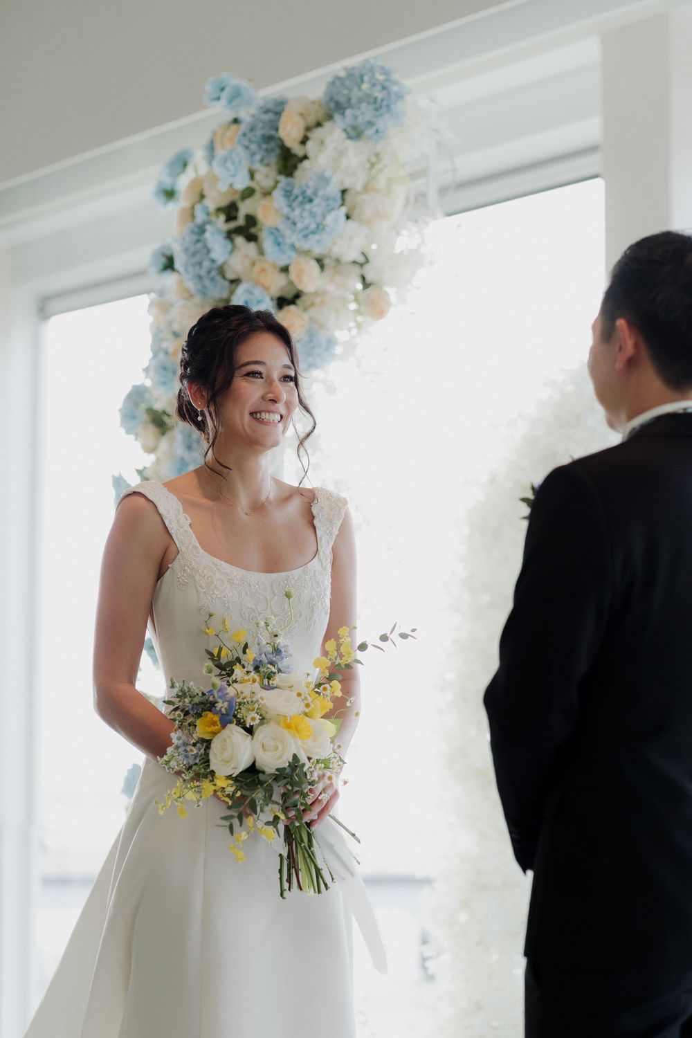 Smiling bride in white gown holds yellow and white bouquet at altar with blue hydrangea floral arch backdrop.