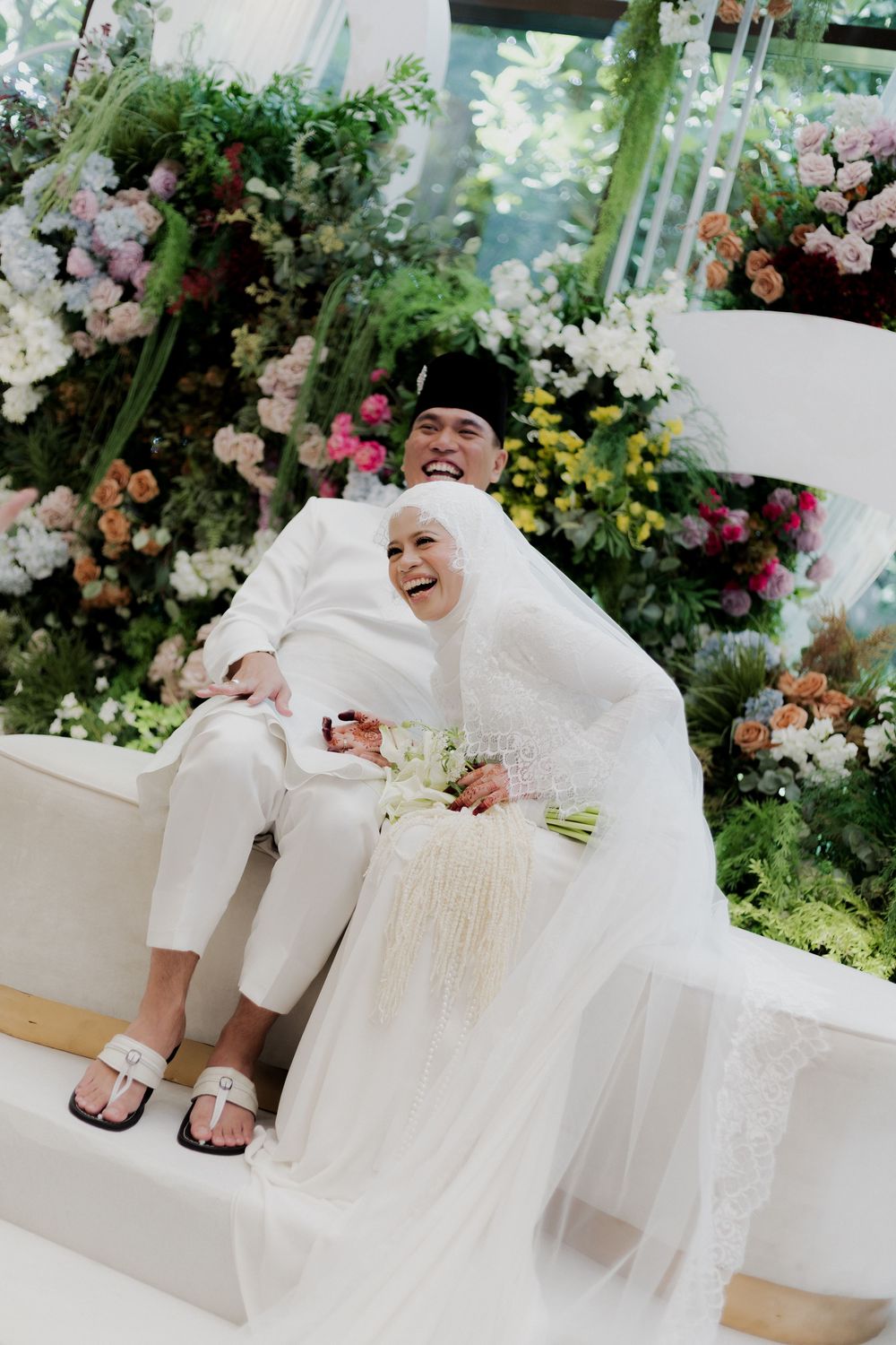 Muslim couple in white wedding attire laughing joyfully, surrounded by lush floral arrangements in an elegant ceremony setting.