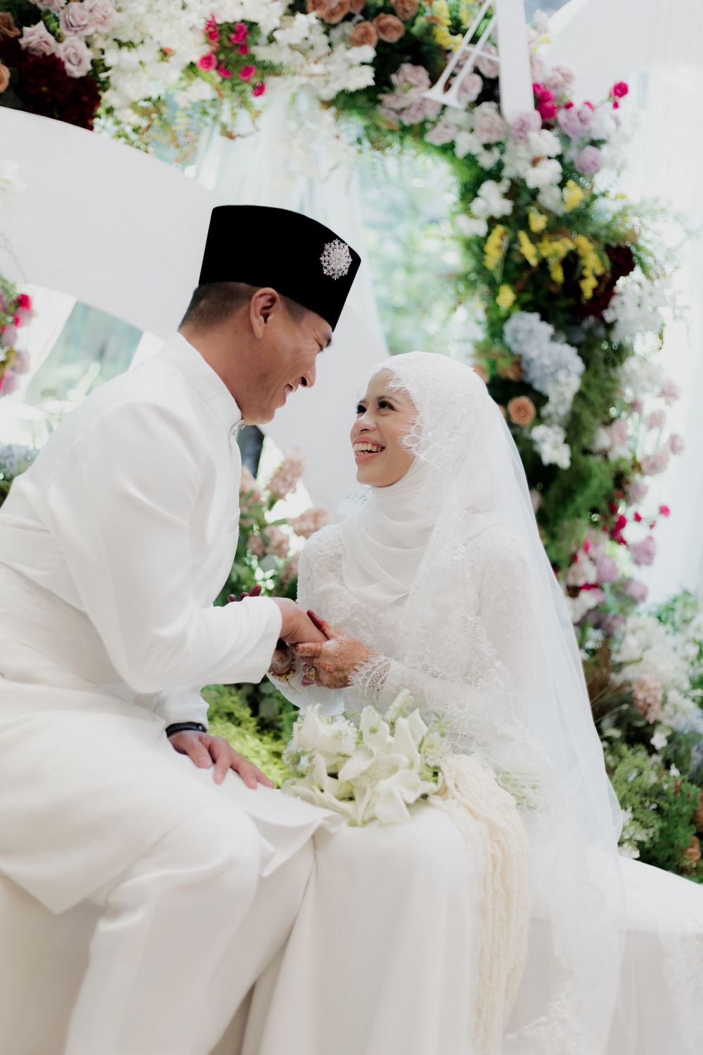 Muslim bride and groom in white attire sharing a tender moment at a floral wedding ceremony.