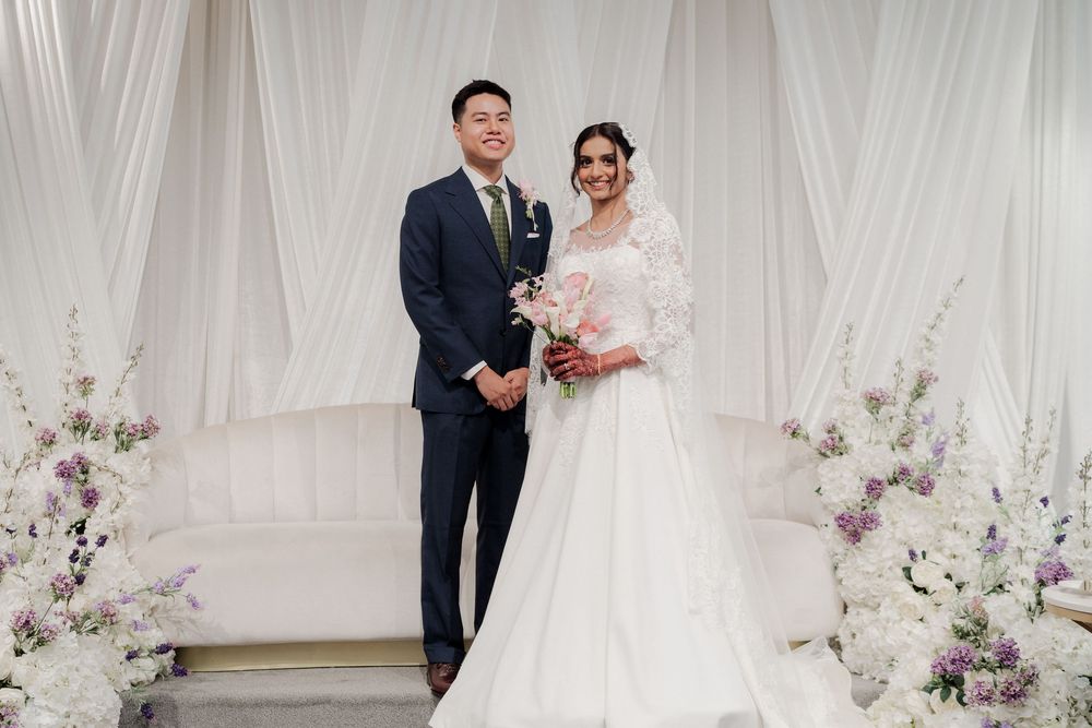 Wedding couple posing against white backdrop with purple floral decorations.
