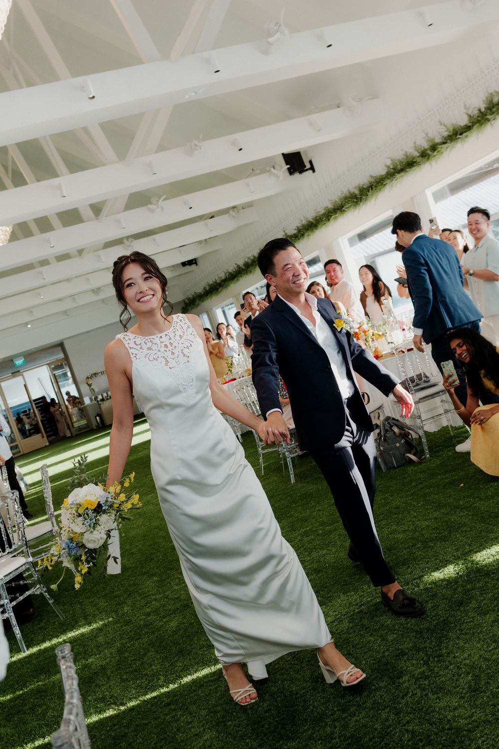 Bride in white gown and groom in navy suit walk happily through wedding venue with green walls and guests.