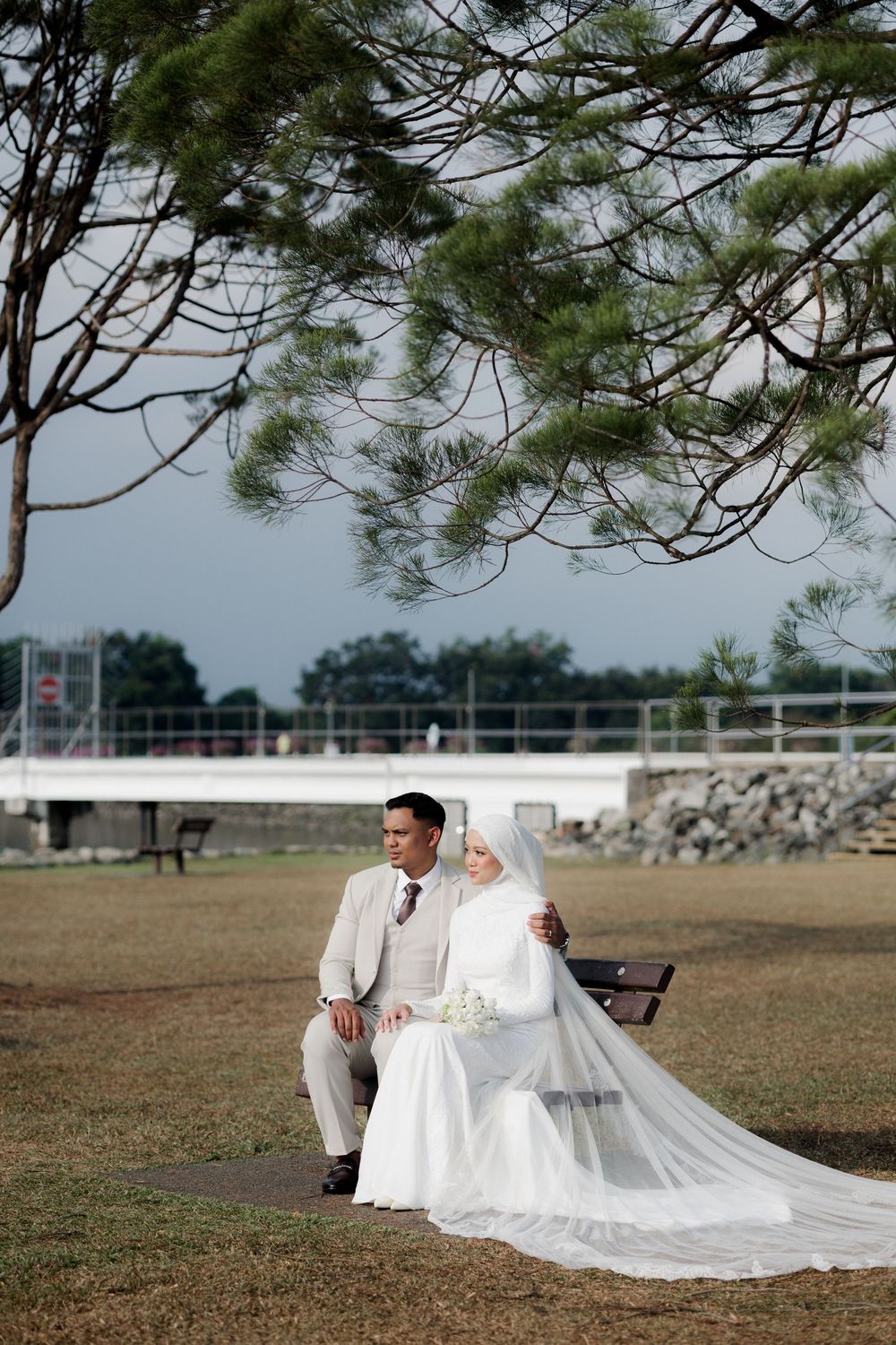 Muslim couple in wedding attire sitting on a park bench, bride in white hijab and gown, groom in beige suit.