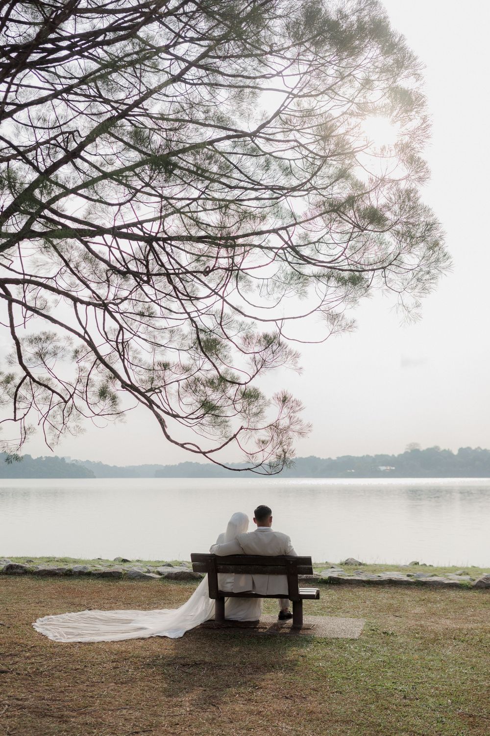Bride and groom sitting on a bench by a serene lake under a large tree, with a flowing white wedding dress train.