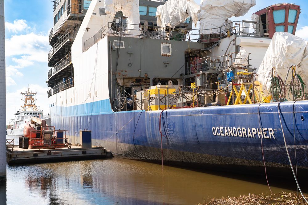 Large cargo ships and vessels docked at a commercial port with industrial equipment visible along the waterfront.