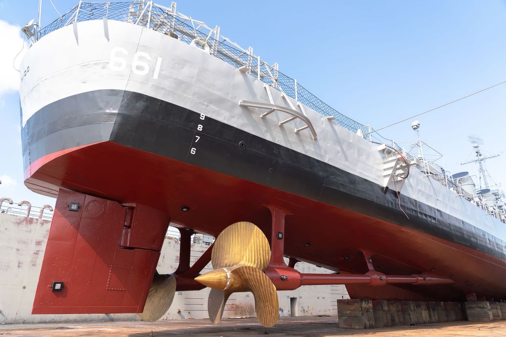 A sleek red and white ship hull with geometric angles sits in dry dock during construction or maintenance work.