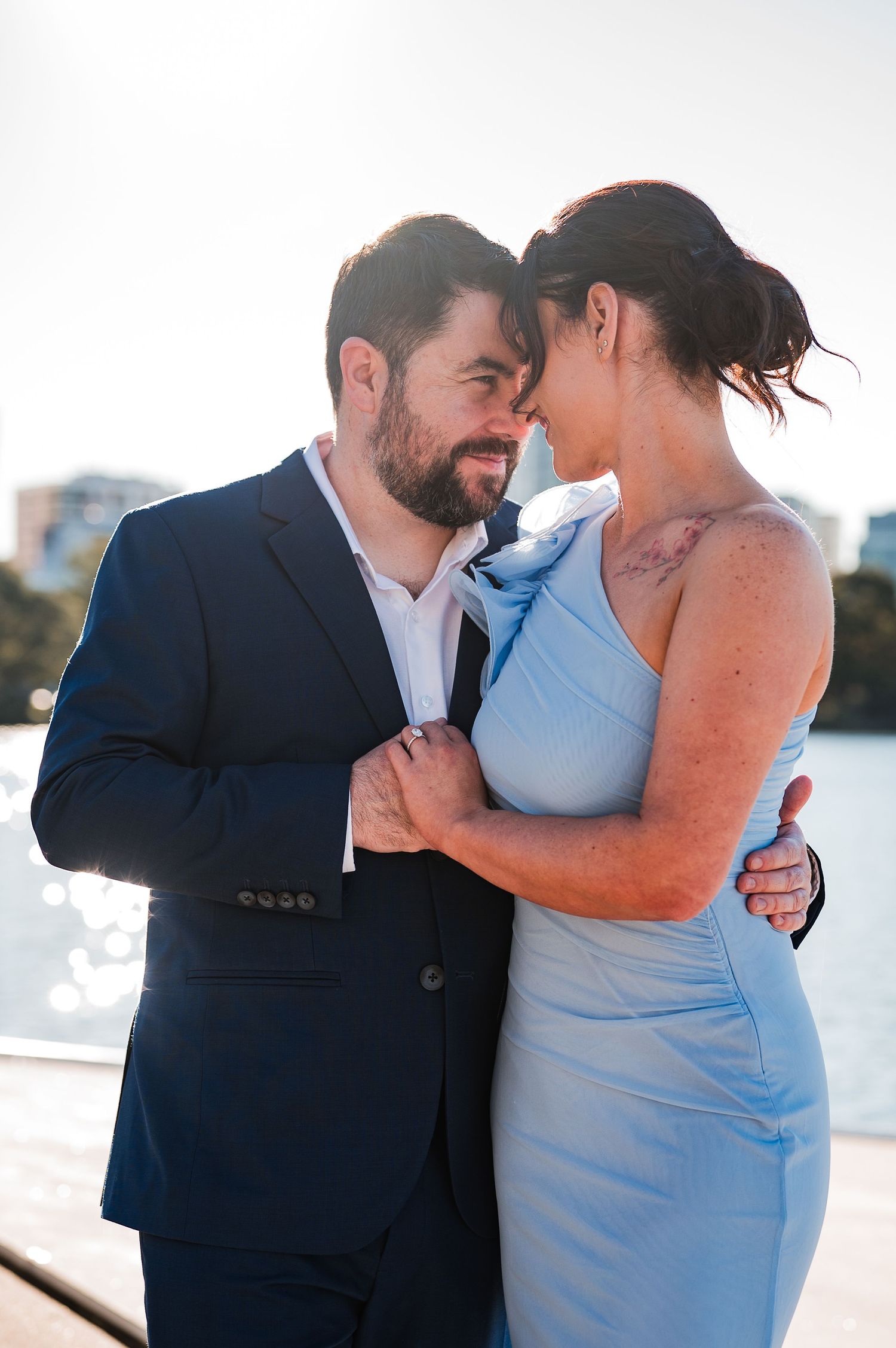 Couple in formal attire sharing a romantic moment by the waterfront, man in navy suit, woman in light blue dress.