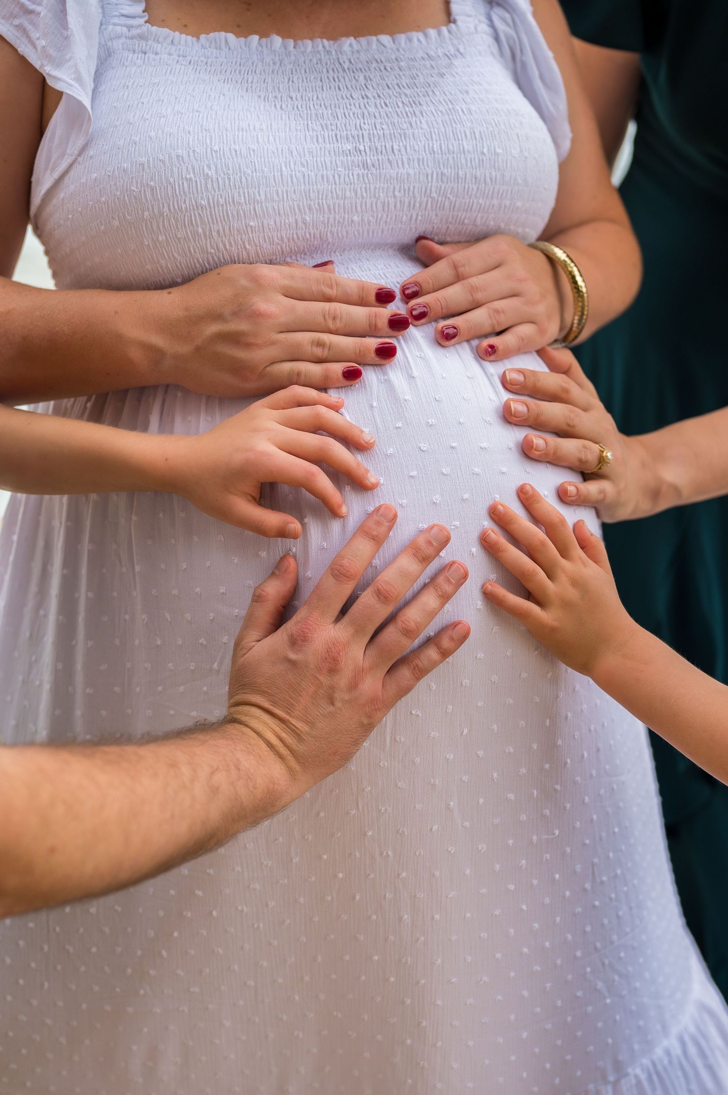 Multiple hands touch a pregnant woman's belly wearing a white dress with red nail polish visible.