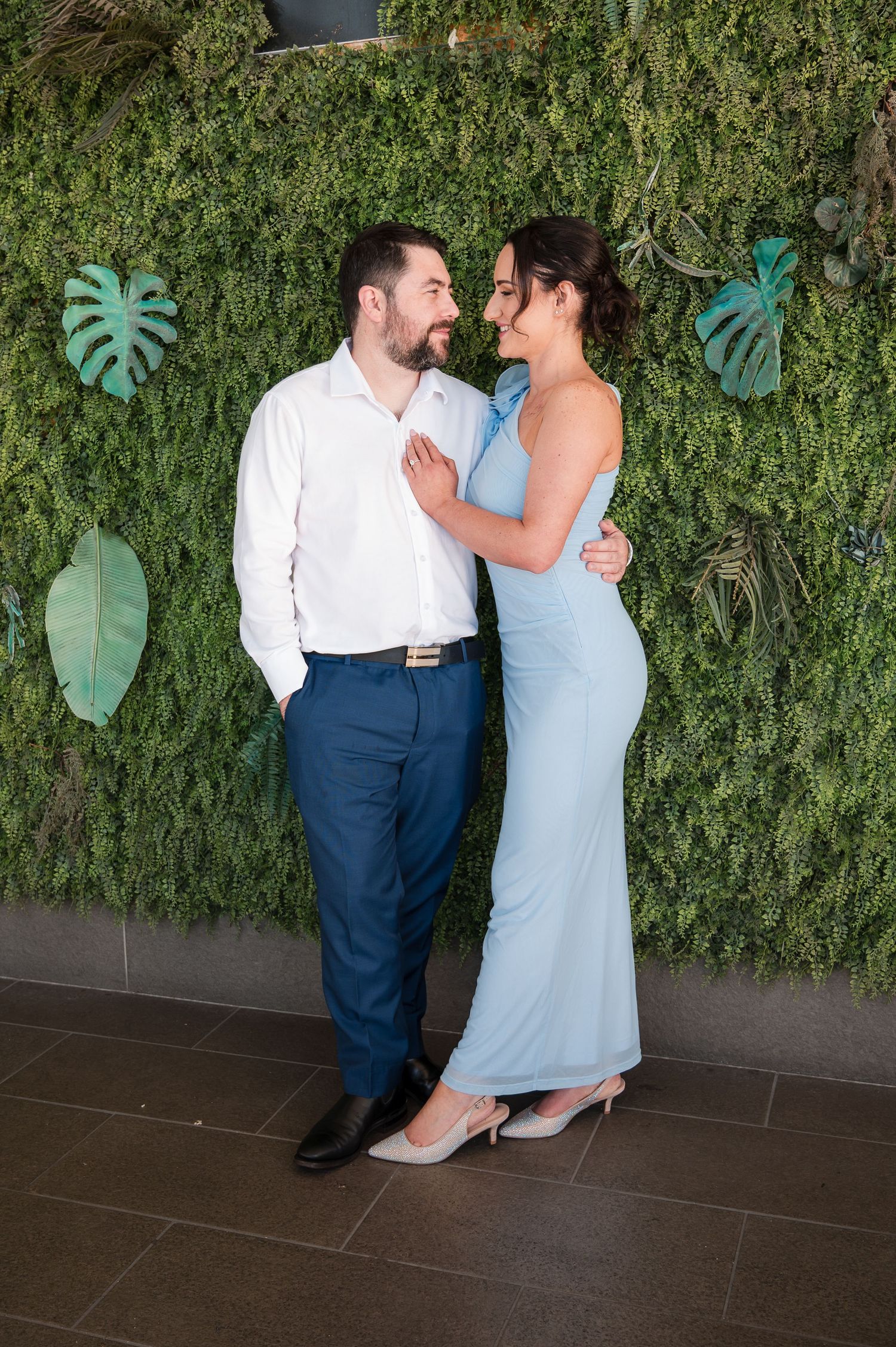 Couple in formal attire smiling at each other in front of a lush green plant wall with tropical leaf decor.