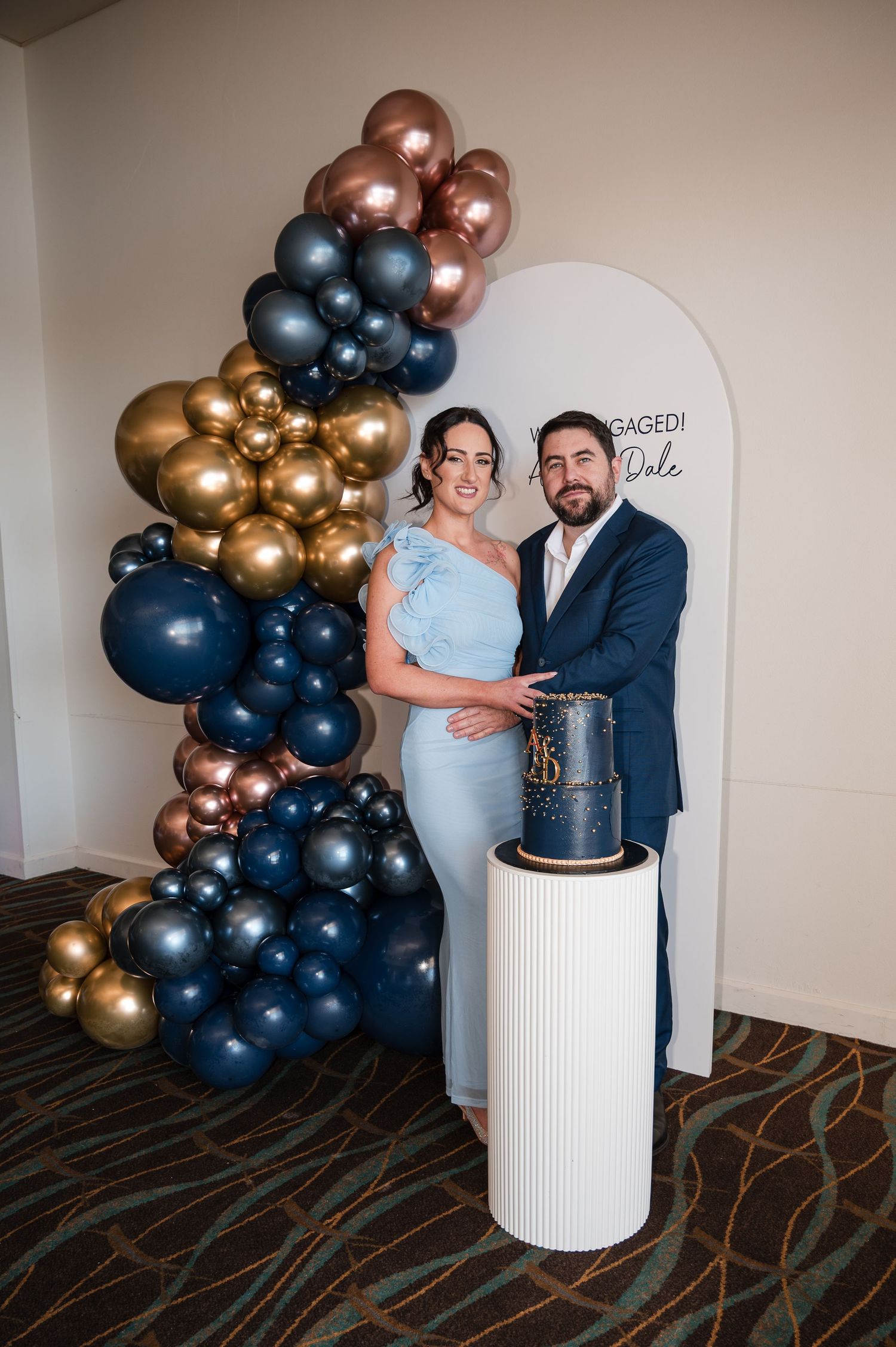 Couple poses by navy, gold, and rose gold balloon arch with elegant cake at engagement party.