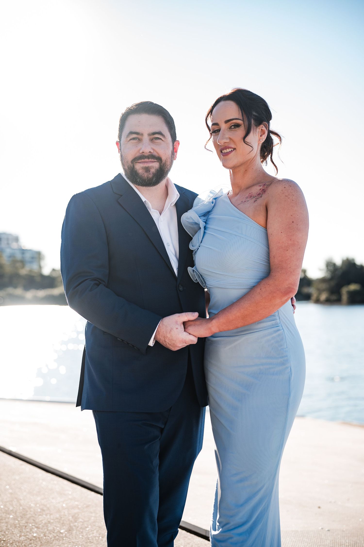 Couple posing by waterfront, man in navy suit and woman in light blue one-shoulder gown, smiling at camera.