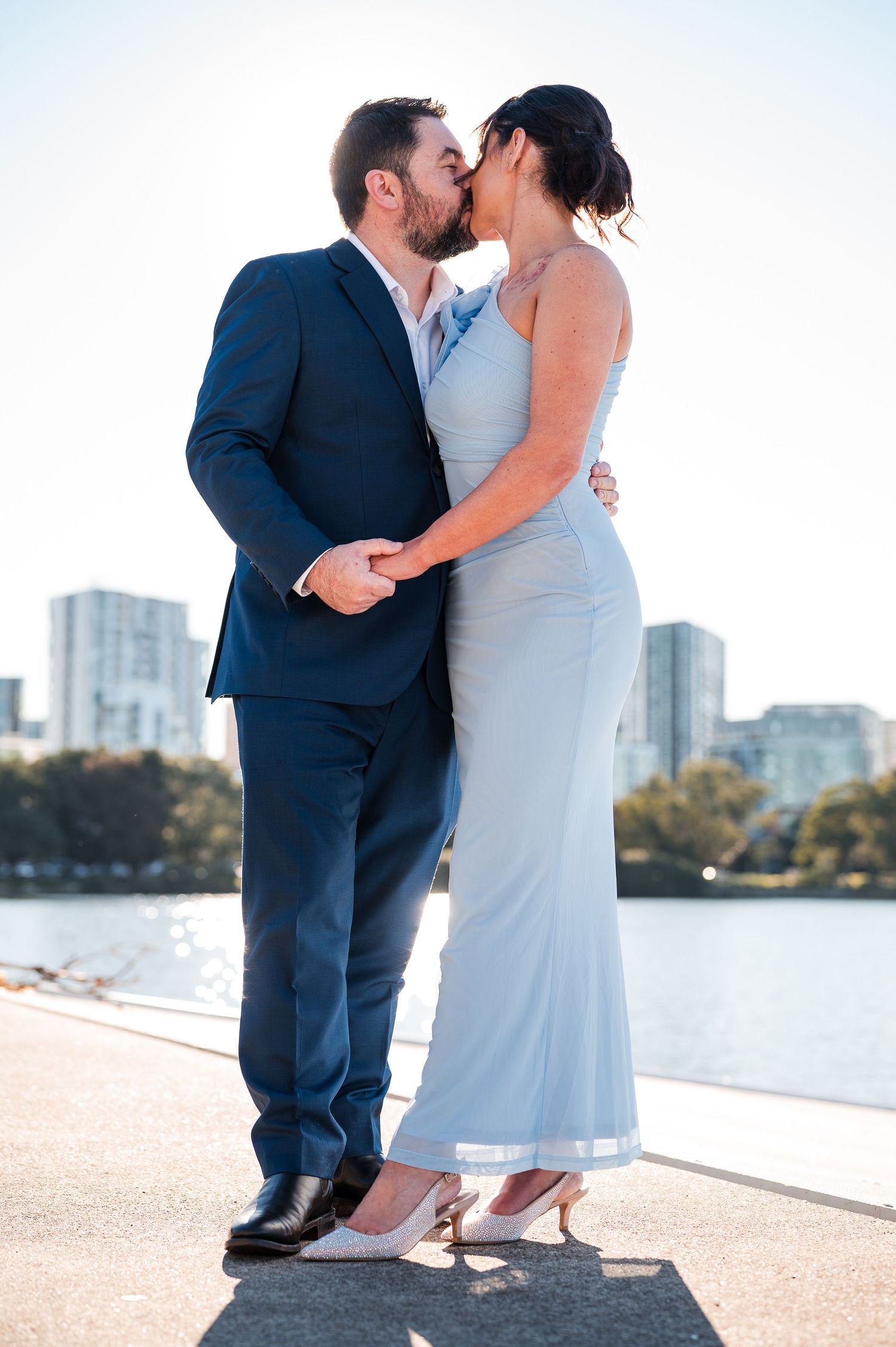 Couple sharing a romantic kiss outdoors near a waterfront, man in navy suit and woman in light blue gown.