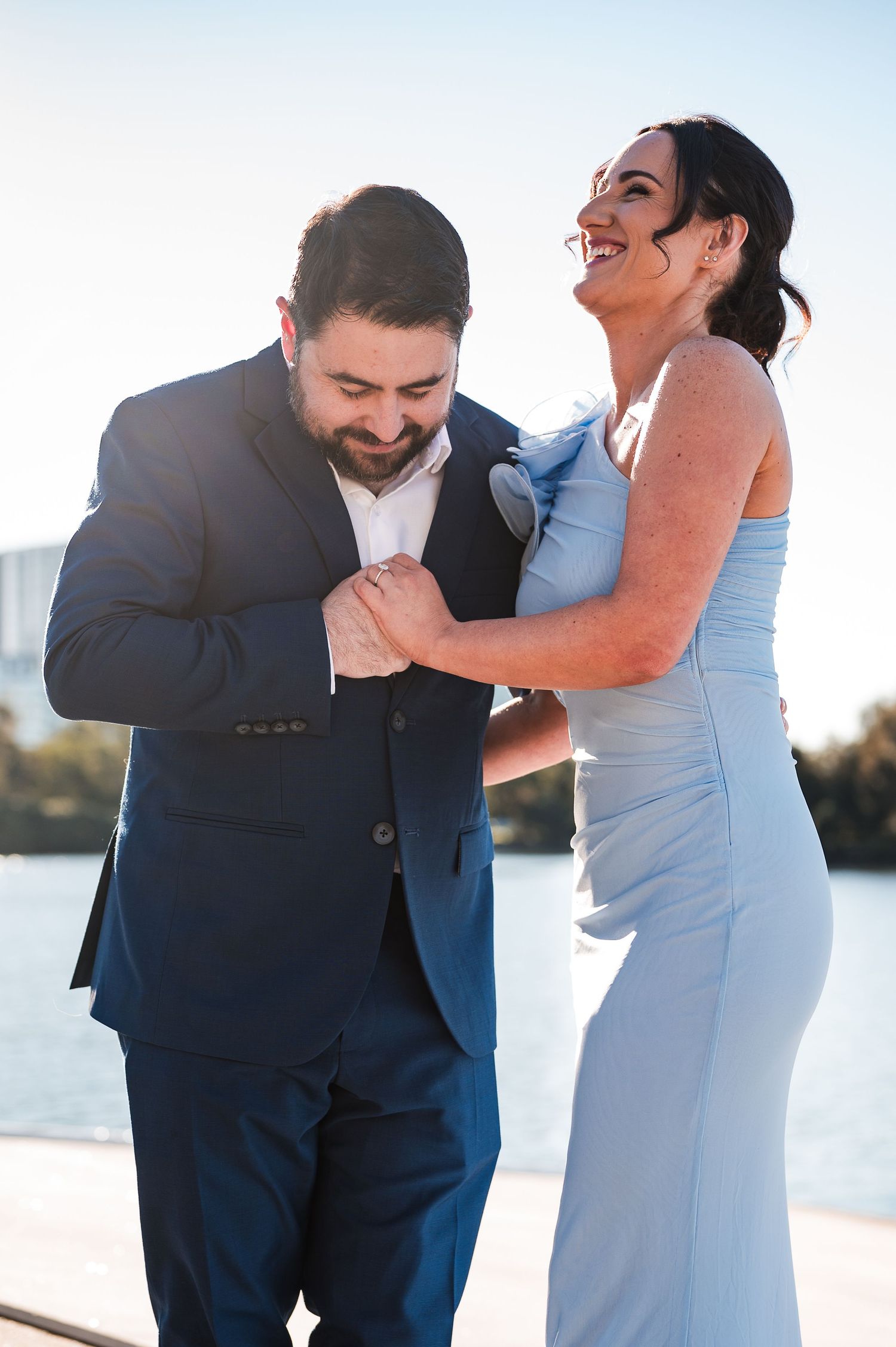 A laughing couple in formal attire, man in navy suit and woman in light blue gown, share a joyful moment outdoors.