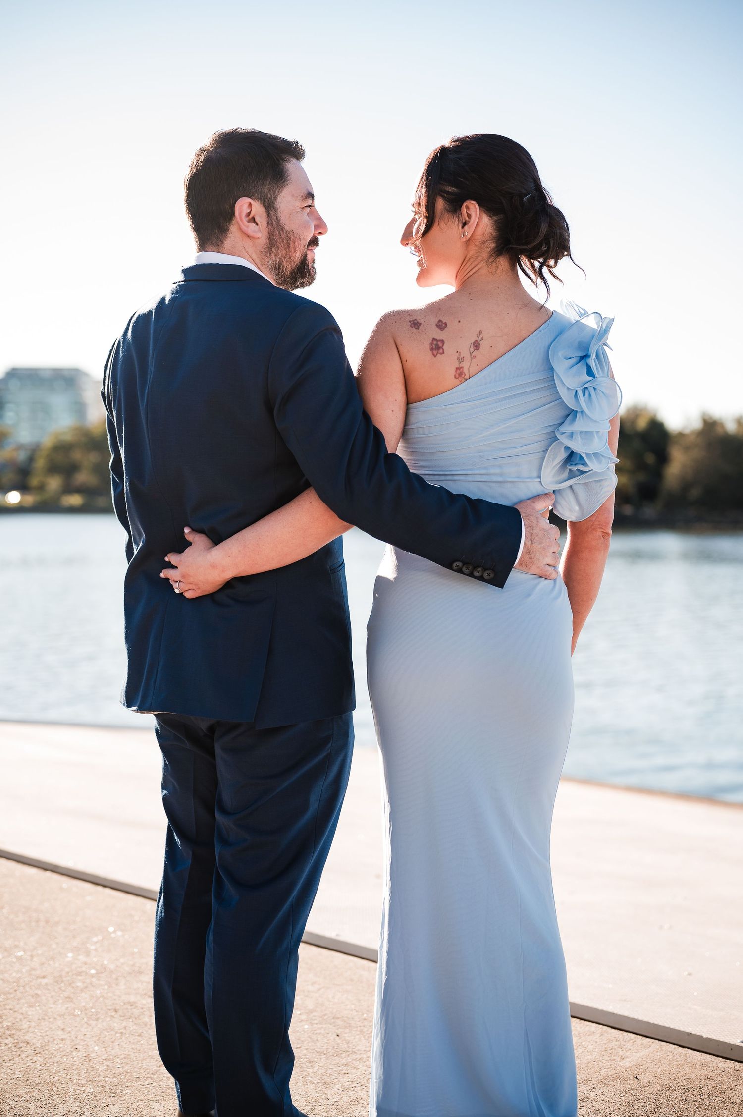 Couple in formal attire sharing a romantic moment by the waterfront, woman in light blue gown with floral tattoo.