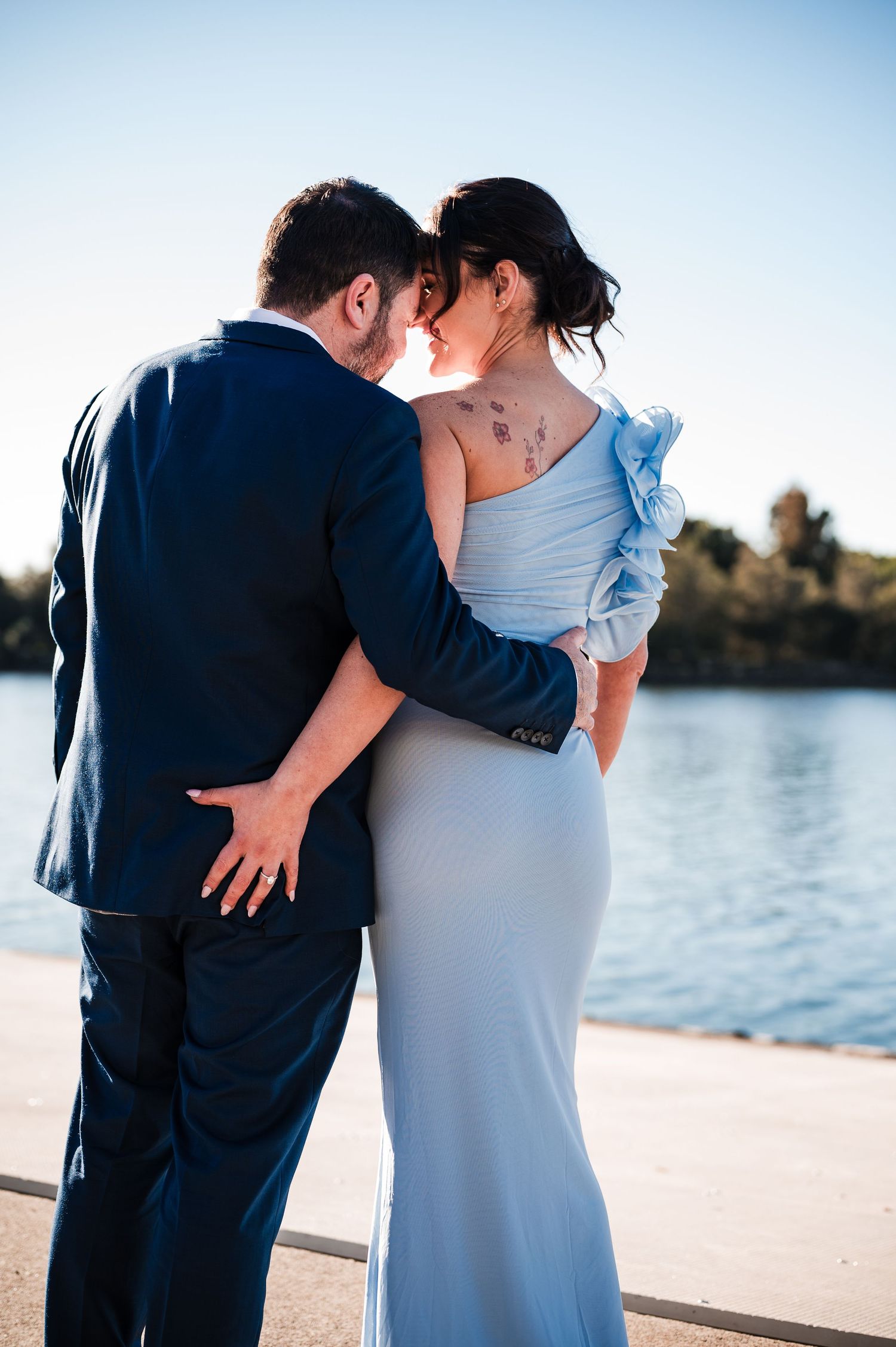 Couple embracing by waterfront, woman in light blue gown with tattoo, man in navy suit, romantic outdoor setting.