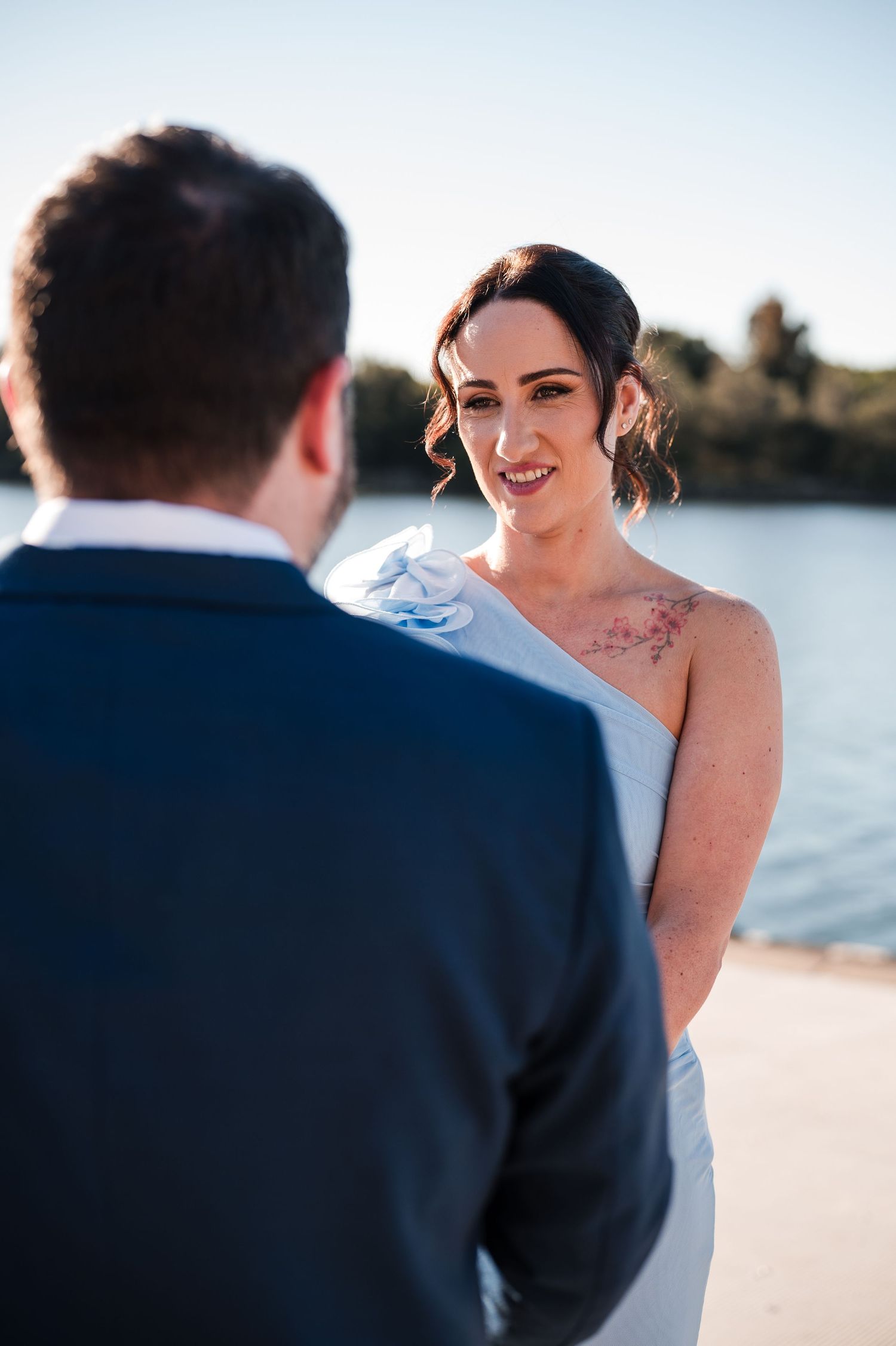 Bride smiling at groom during outdoor beach wedding ceremony, wearing white gown with floral shoulder tattoo.