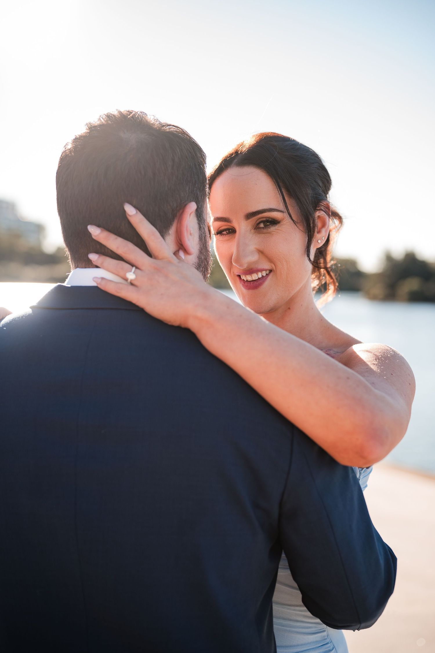 Bride smiling over groom's shoulder during an embrace, wearing wedding ring, with waterfront backdrop.
