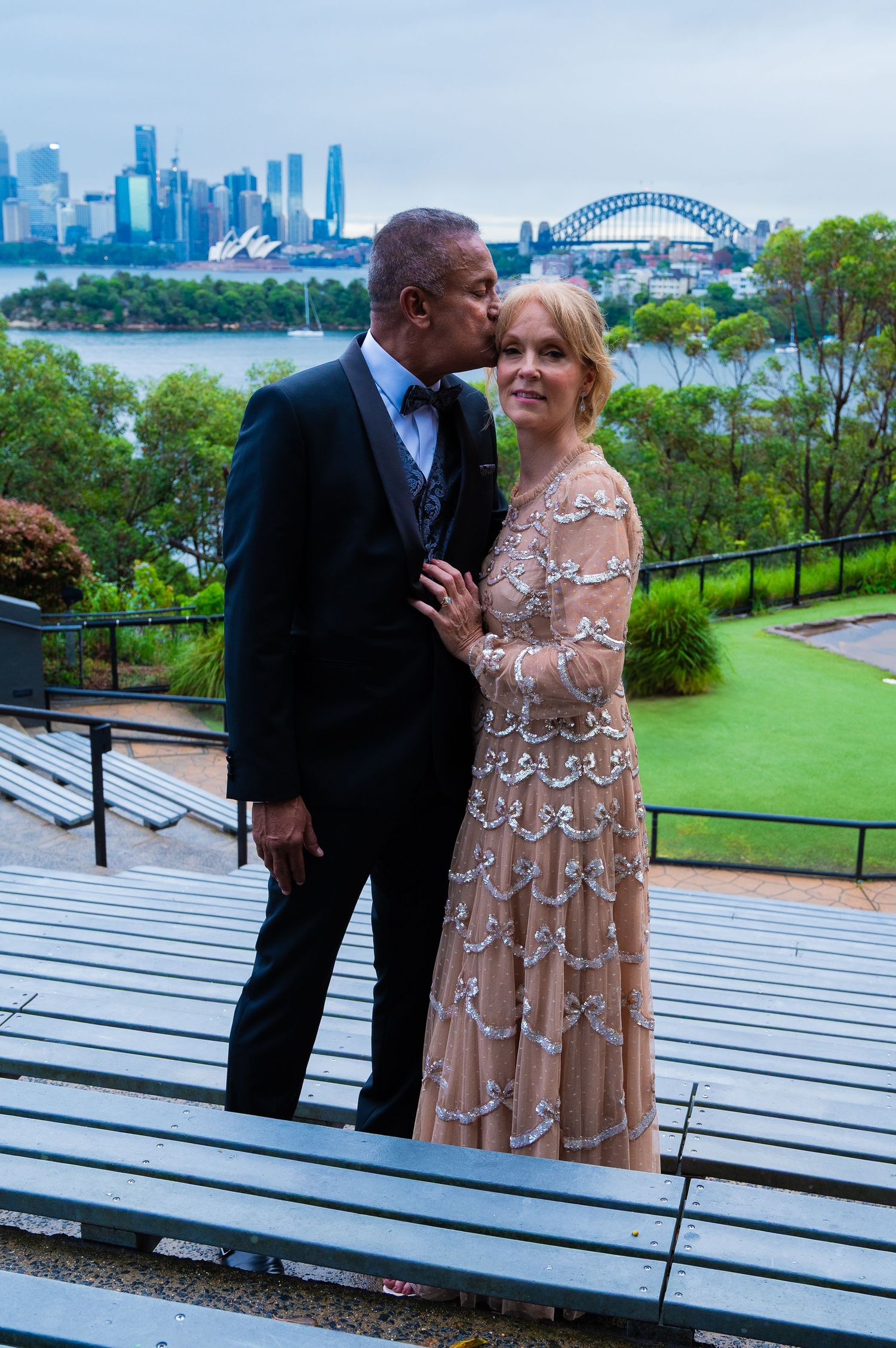 A couple shares romantic moments at a scenic overlook with Sydney Harbour Bridge and city skyline in the background.