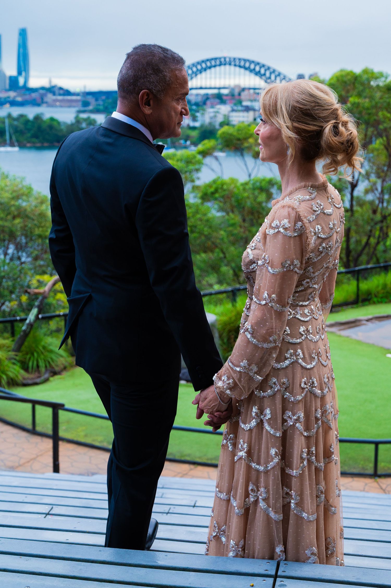 Two people wearing formal attire stand together on a deck overlooking Sydney Harbor Bridge and city skyline.
