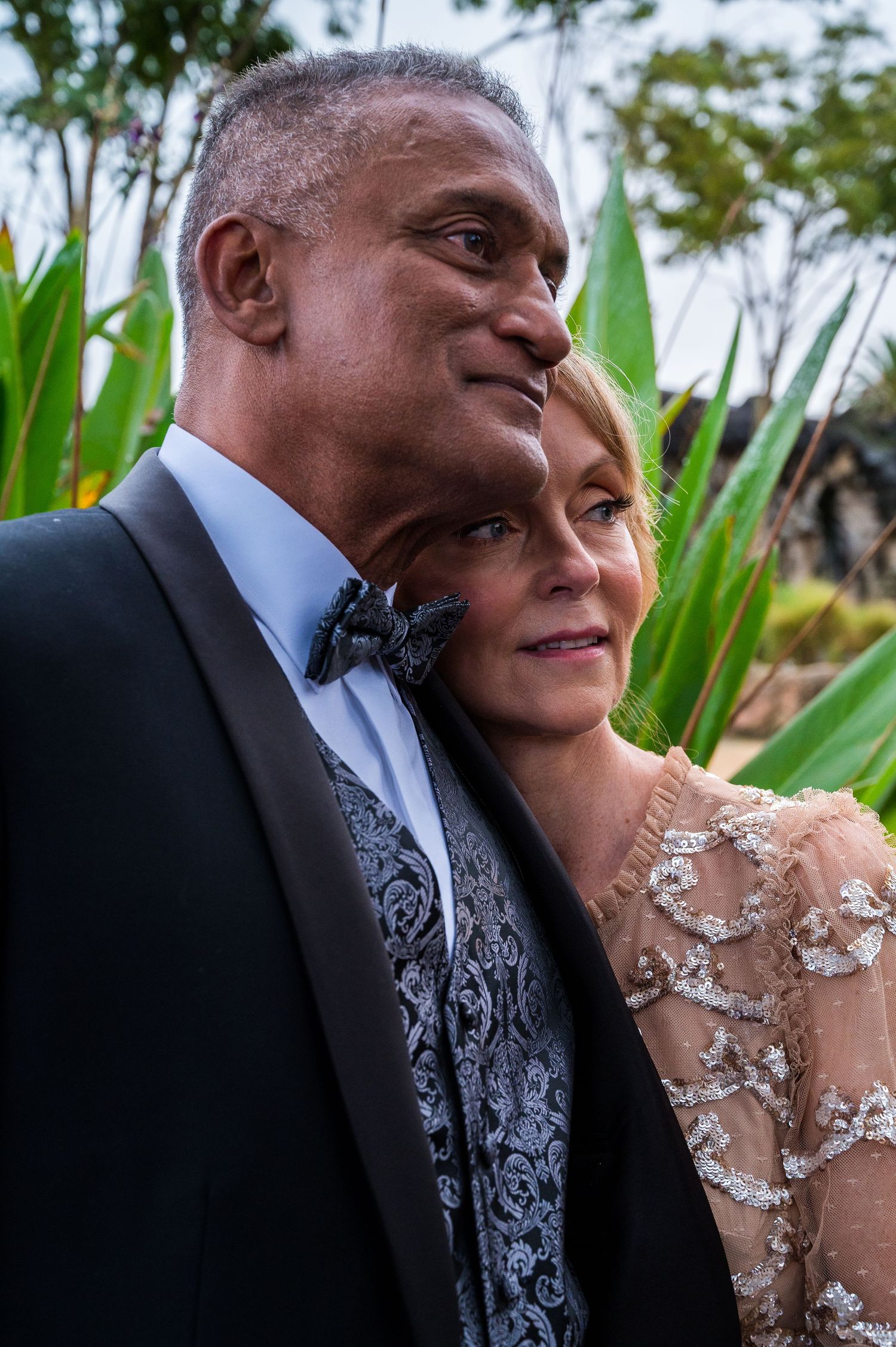 A couple in elegant wedding attire gazes into the distance, with tropical greenery in the background.