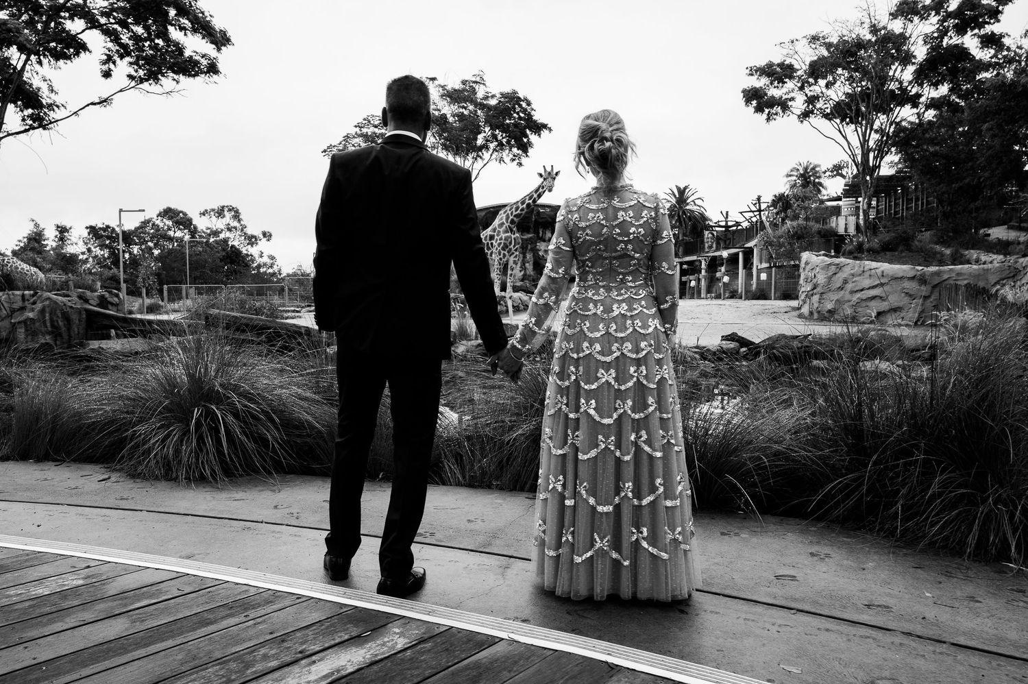 A couple stands together on a wooden deck surrounded by coastal landscaping, captured in black and white.