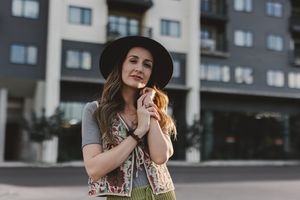 Stylish young woman in black wide-brim hat and floral vest posing outdoors near modern apartment building.
