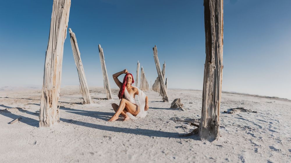 Woman in white bikini sitting among weathered wooden posts on a vast white salt flat under a clear blue sky.