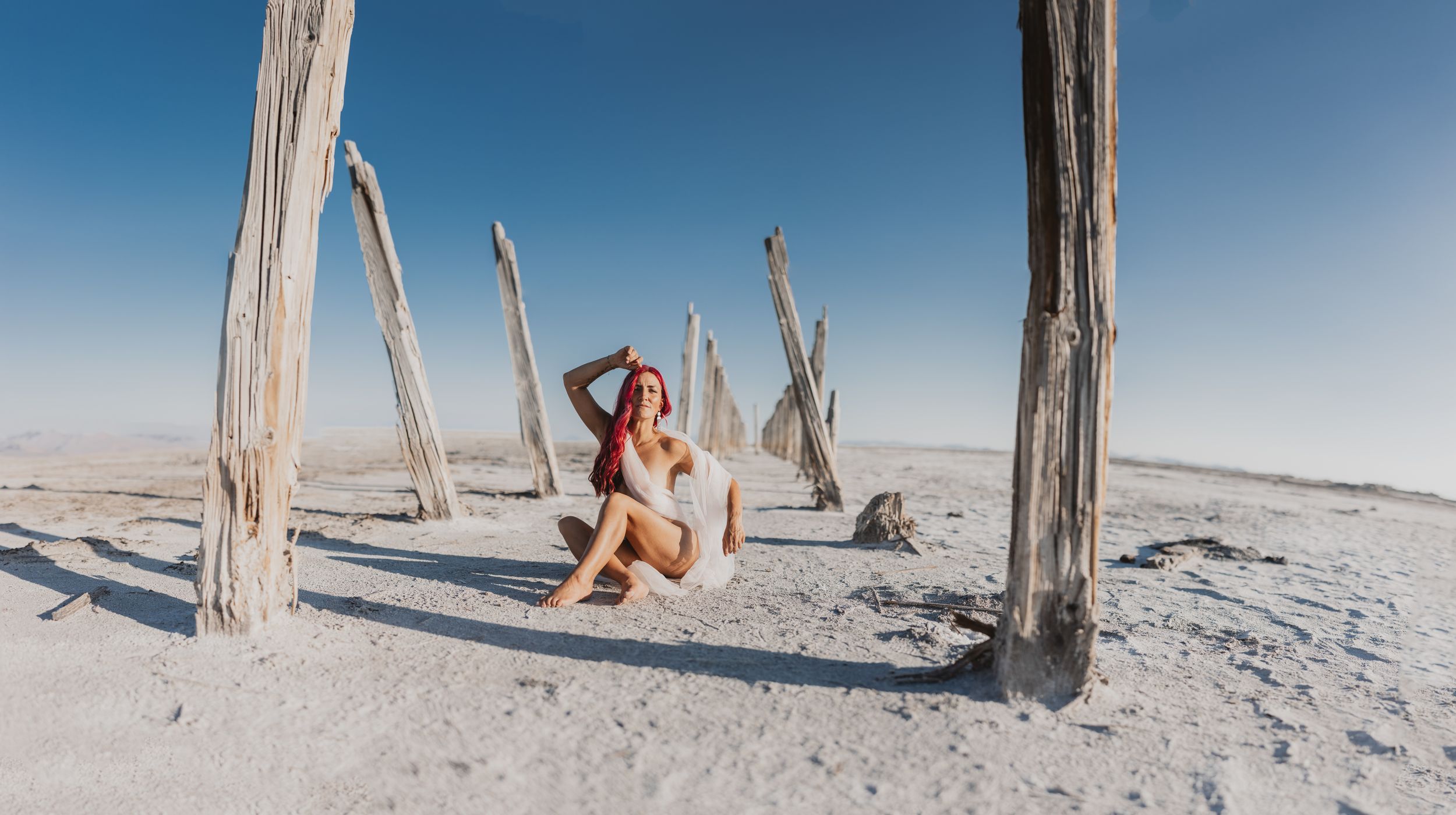 Woman in white bikini sitting among weathered wooden posts on a vast white salt flat under a clear blue sky.