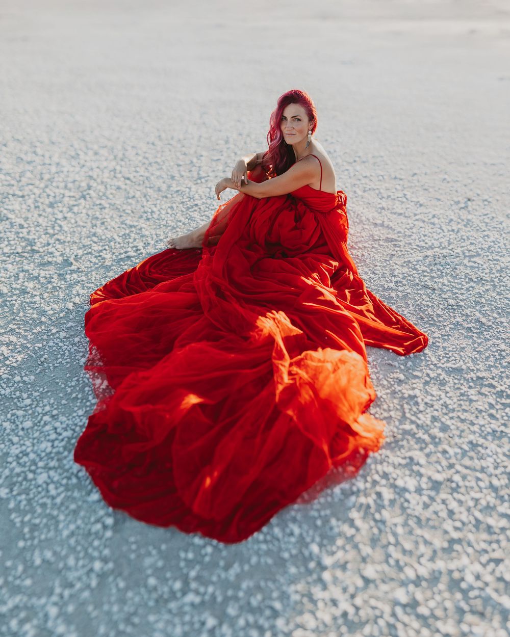 Woman in flowing red gown sitting on white salt flats, her red hair matching the dramatic billowing dress.