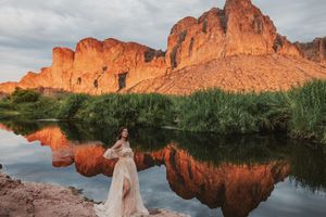 Woman in flowing gown poses by reflective desert river with glowing red rock mountains at sunset.