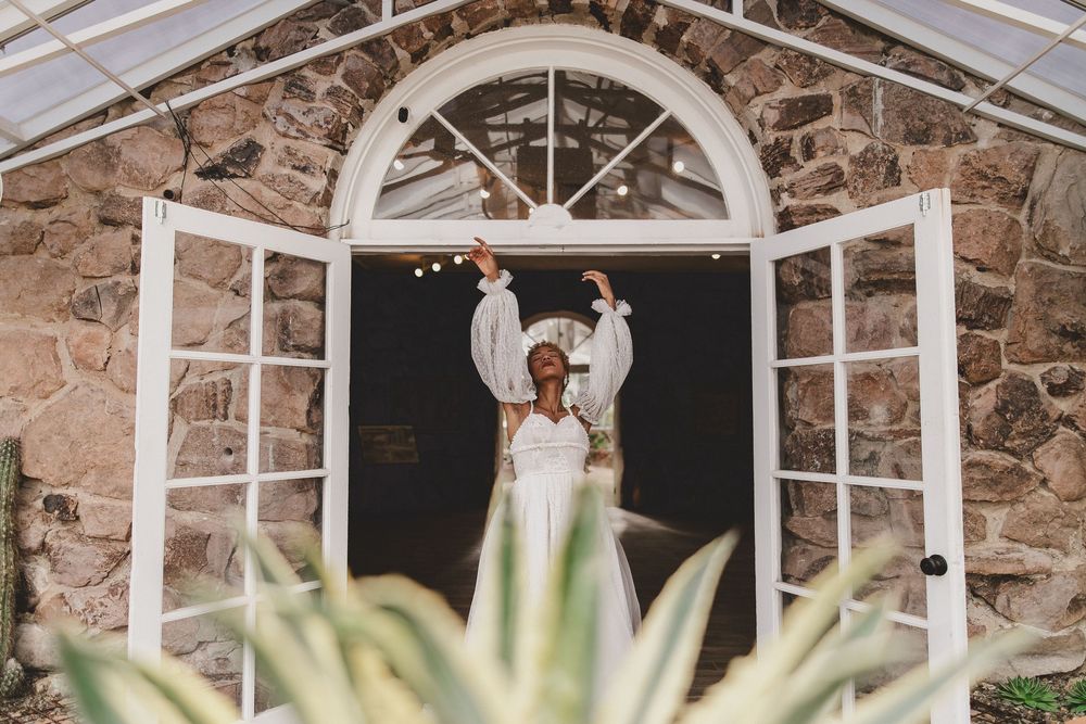 Bride in white dress raises arms joyfully in arched doorway of rustic stone building with fairy lights.