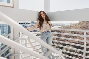 Young woman in casual jeans and beige top posing on a bright white balcony staircase with city views.