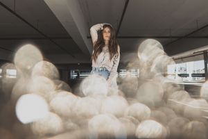 Young woman posing in parking garage surrounded by dreamy bokeh light orbs, wearing denim shorts and gray top.
