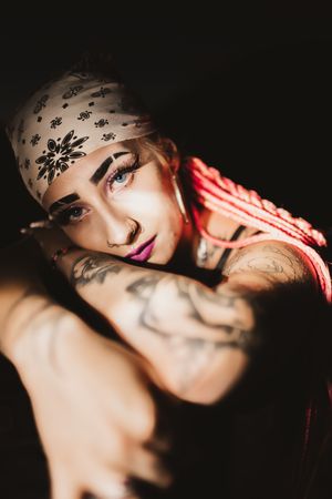 Tattooed person wearing a white bandana headband with bold makeup including purple lips poses against dark background.