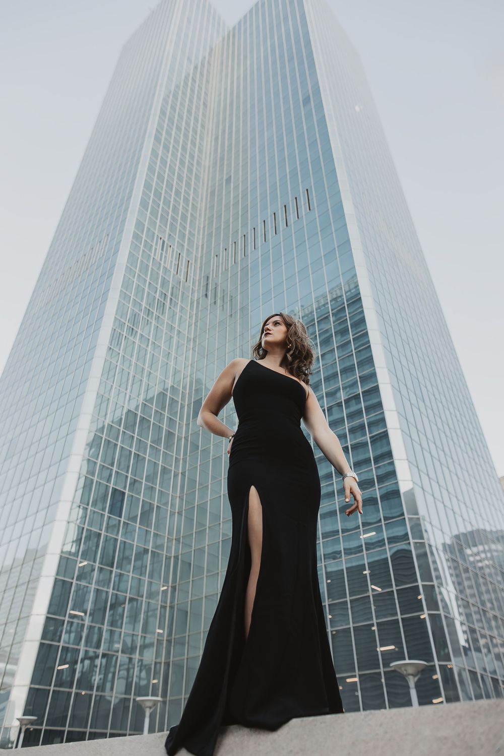 Woman in elegant black slit gown poses confidently in front of modern glass skyscraper in urban setting.