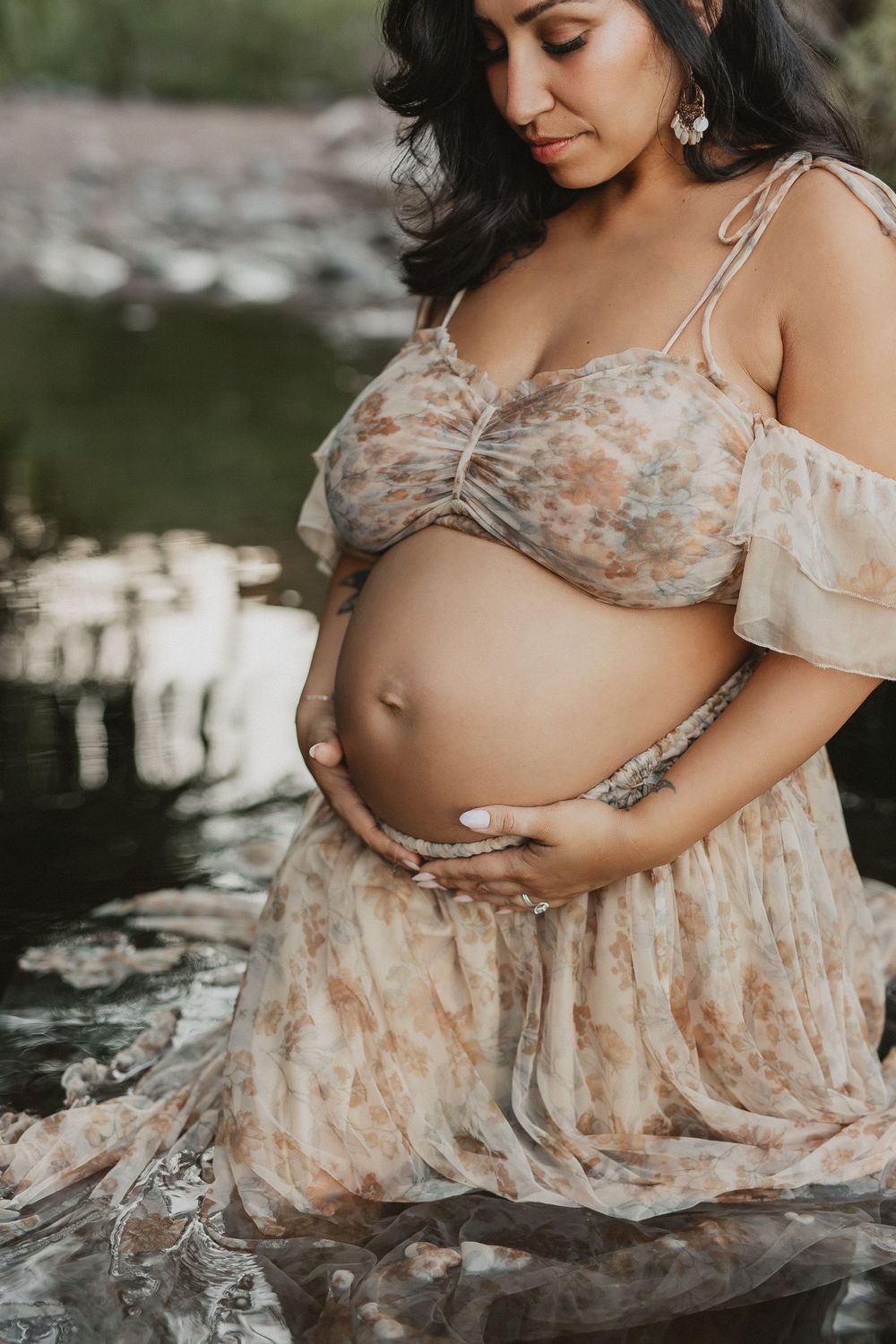 Pregnant woman in floral dress cradling baby bump near a serene river during an outdoor maternity photoshoot.
