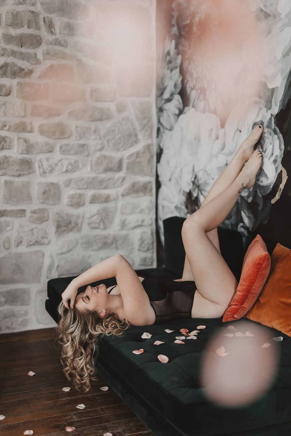 Woman with blonde curly hair posing on a dark velvet sofa with orange pillows in a stylish studio setting.