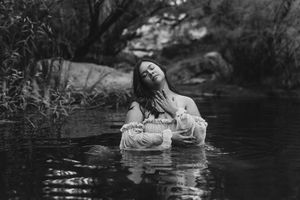 Black and white photo of a woman in white dress sitting in a serene river, head tilted back with eyes closed.