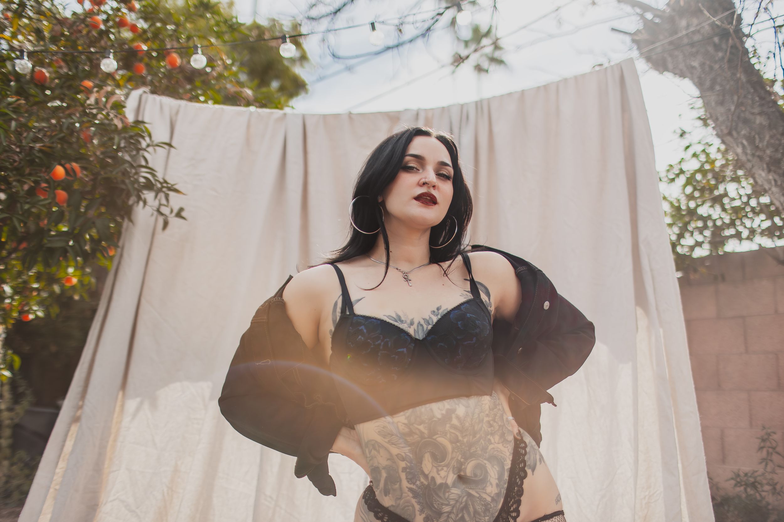 Woman in black lace lingerie and gloves poses outdoors near white draped fabric and orange tree.