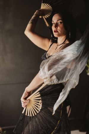 A woman in a black dress poses dramatically with two decorative hand fans in a dimly lit studio setting.