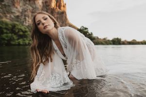 Woman in white lace dress wading in a river near rocky cliffs, looking up gracefully.
