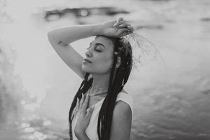 Black and white photo of a young woman with braids posing gracefully near water, hand raised to her wet hair.