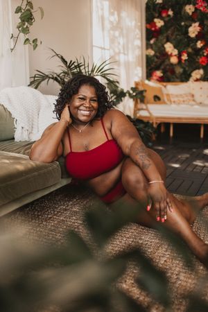 Smiling plus-size Black woman in red bikini sitting on floor beside sofa, surrounded by indoor plants and flowers.