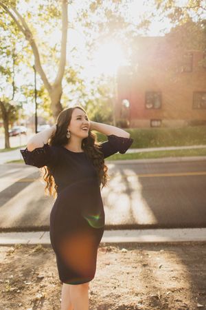 Woman in black dress posing outdoors with sun flare, long wavy hair, smiling joyfully on a sunny street.