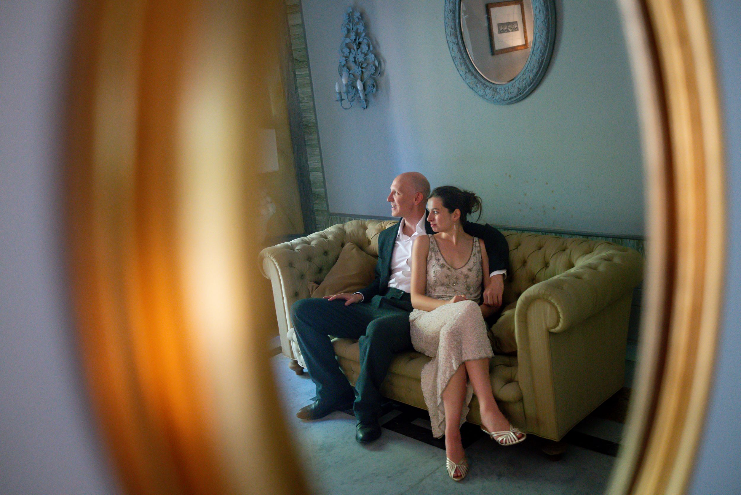 Couple reflected in ornate gold mirror, sitting intimately on velvet sofa in elegant blue room.