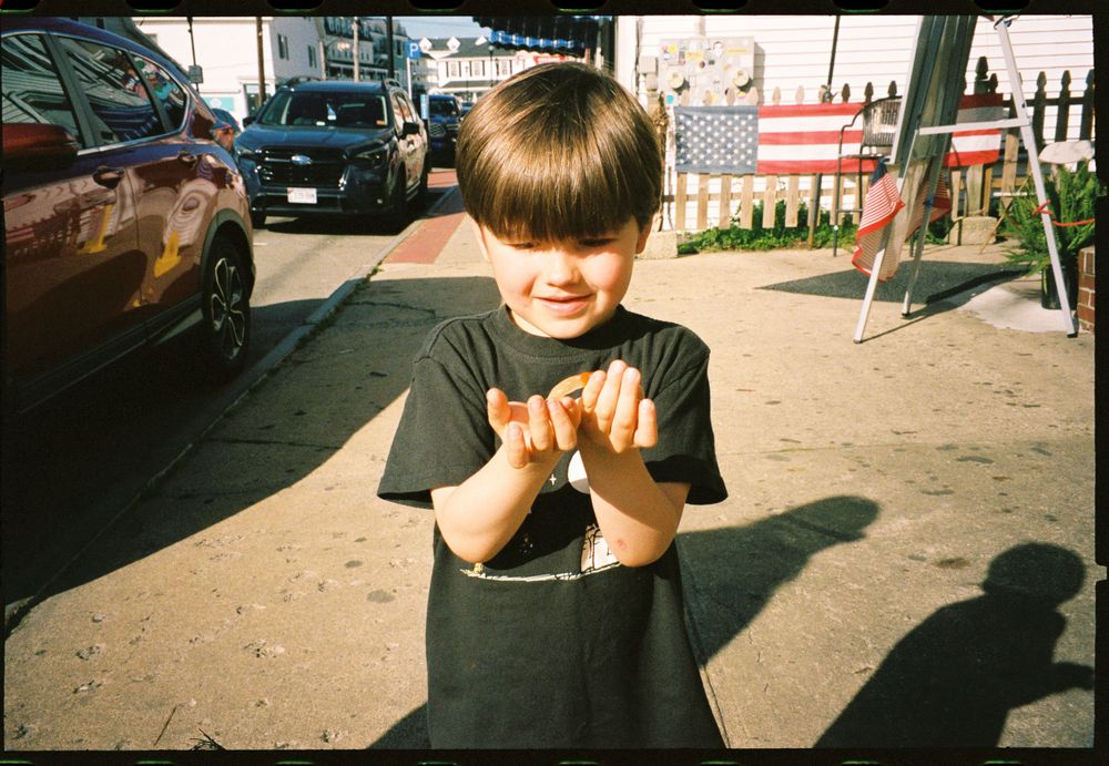 Child in black shirt smiling while standing on a sidewalk near parked cars.