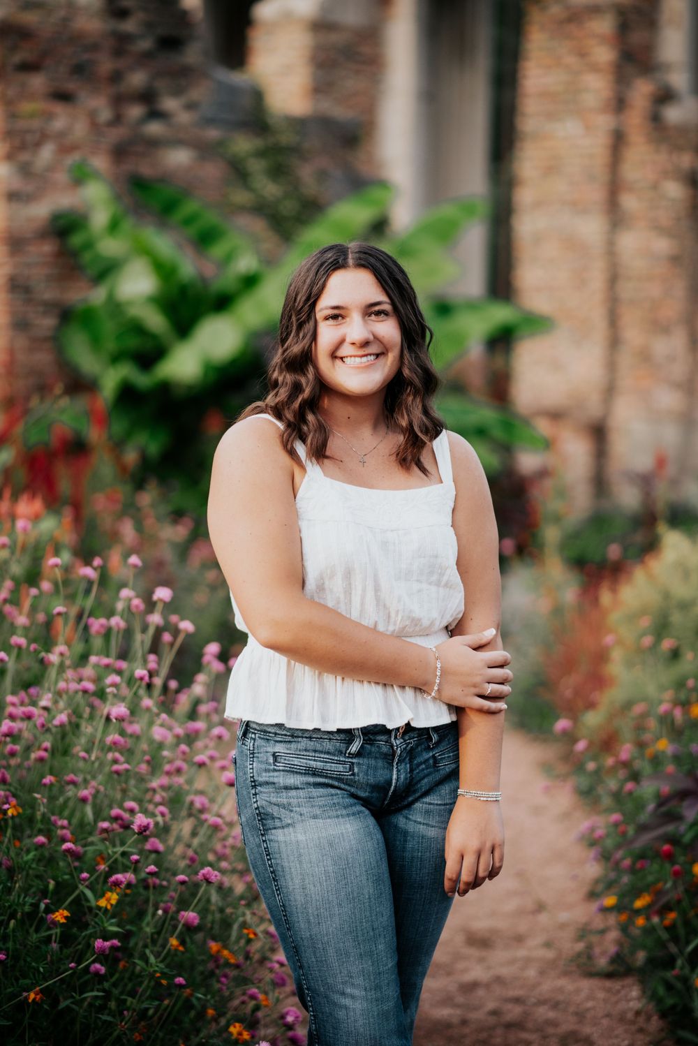 Senior portrait in a white top and jeans standing among colorful garden flowers.