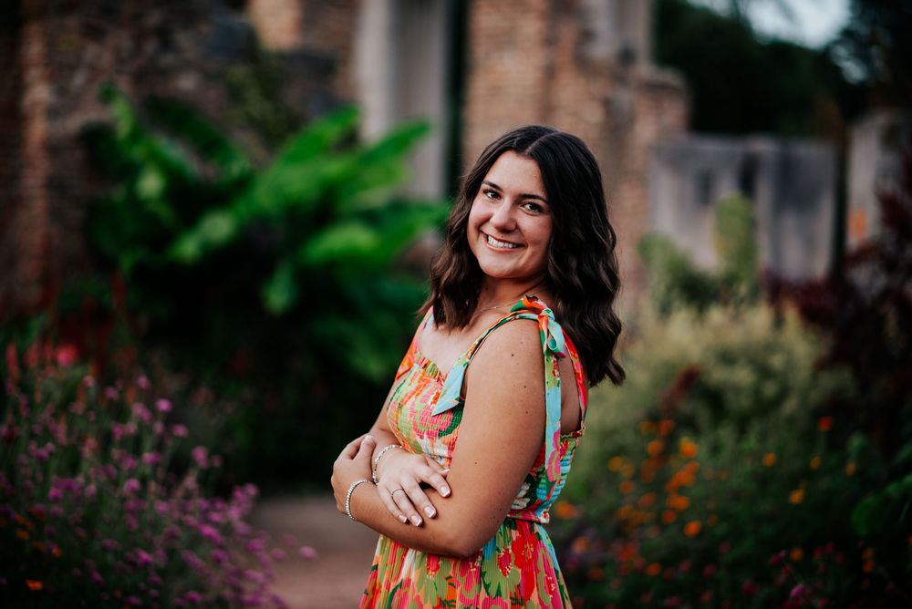 Senior portrait in a floral dress posing in a garden with brick wall background.